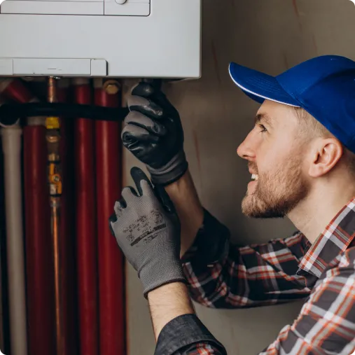 Man in plaid shirt and blue cap working on a boiler, adjusting controls with gloved hands.