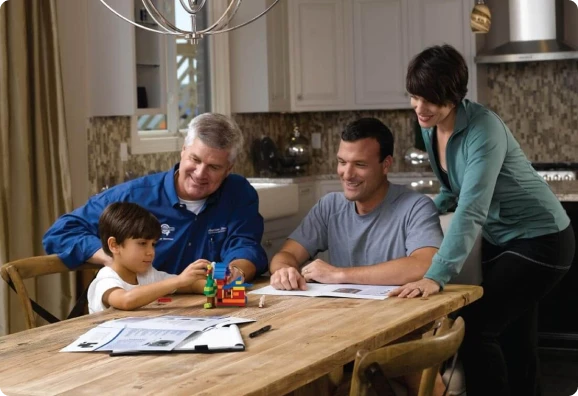 Three adults and a child sit around a wooden table with papers and toys in a kitchen.